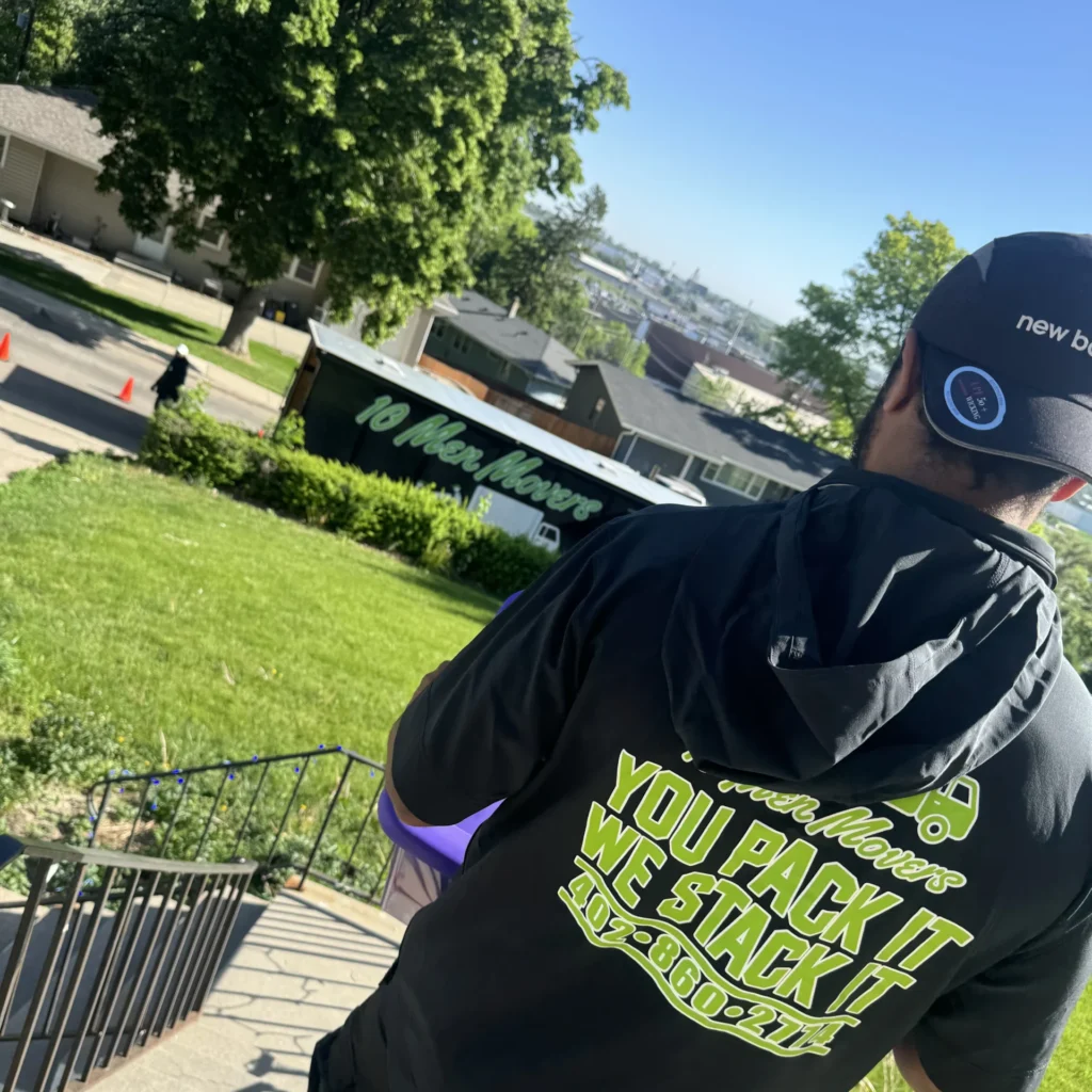 Omaha mover in branded attire carrying a box, with a 10 Men Movers truck in the background, emphasizing local moving services.